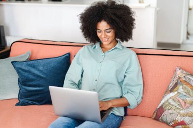 Clever female freelancer using laptop for remote work from home. Charming black woman web surfing, sending and answering emails, developing software while sitting at the sofa