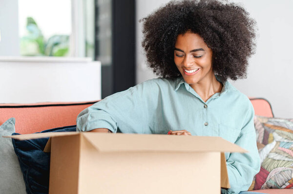 Happy with delivery satisfied female customer unpacking cardboard box, excited African-American woman glad to get a parcel, order online, overjoyed girl at home