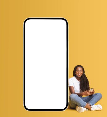 Afrika kökenli Amerikalı bir kadın boş ekranlı büyük bir akıllı telefonun yanında oturuyor, elinde akıllı telefon, sosyal ağlarda sohbet etmekten, mesajlaşmaktan ve gülümsemekten hoşlanıyor,