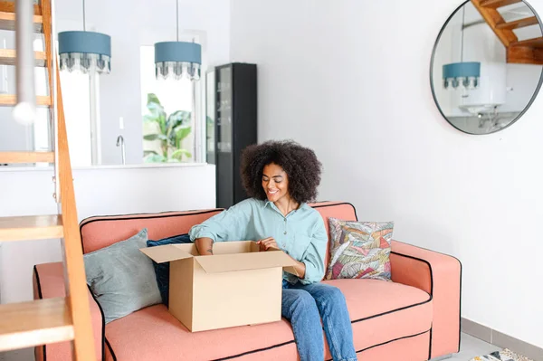 Happy multiracial woman is looking at her new parcels, sitting on the couch, and unpacking. Smiling african-american lady opening online store order in the postal delivery shipping box