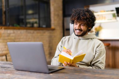 Akıllı günlük giyimli yakışıklı genç adam online dersleri ve webinarları izlemek için dizüstü bilgisayar kullanıyor. O, çağdaş ev ofisinde otururken, deftere notlar alıyor.