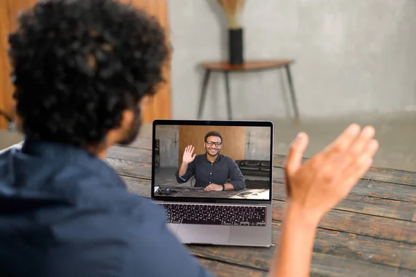Close-up of Pakistani man with curly hair using app for distance video communication with coworker, friend, meeting online, looking and waving at laptop desktop with male profile