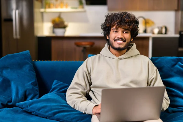 Cheerful Indian Man Wearing Casual Hoodie Sitting Sofa Using Laptop — 스톡 사진