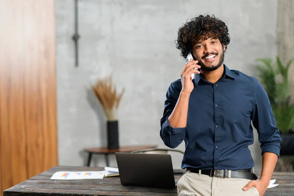 Happy inspired Indian male small business owner talking on the smartphone while standing in modern loft office. Smiling successful contemporary man manager in casual wear holding phone call,copy space