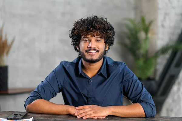 Portrait of happy Indian man sitting at the office desk and looking at the camera, involved in video meeting. Webcam view of smart eastern male in blue shirt