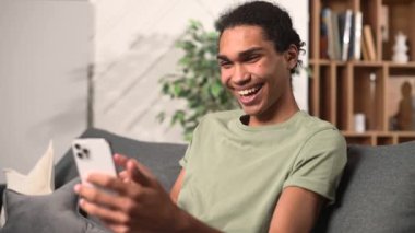 Pozitif genç çok ırklı adam gri koltukta oturuyor, akıllı telefonu tutuyor ve ekrana bakıyor, merhaba diyor, video bağlantısı ile arkadaşlarıyla konuşuyor, video araması yapmak için mobil uygulama kullanıyor.