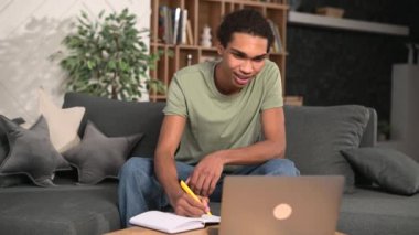 Çok ırklı bir öğrenci, günlük tişört giyiyor, kanepede oturuyor ve ev bilgisayarındaki bilgisayarı izliyor, öğretmenle video görüşmesinde notlar alıyor.