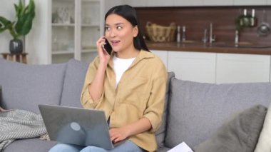 Gülümseyen Asyalı kadın akıllı telefonuyla evde dizüstü bilgisayar kullanıyor, serbest çalışan kadın müşterilerle konuşuyor ve not defterindeki programı kontrol ediyor.