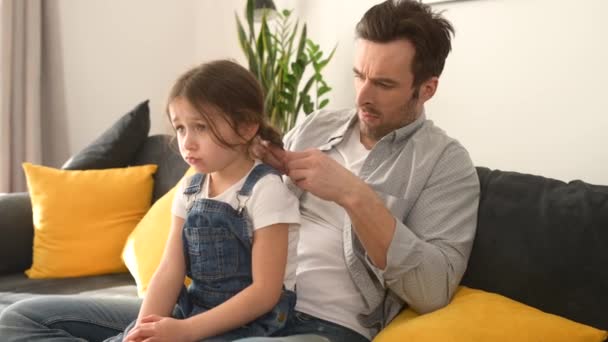 可爱的父亲在舒适的客厅沙发上给他年幼的女儿做发型 在家里做单亲和伤心的孩子 父亲编织辫子 给女儿做头发 — 图库视频影像