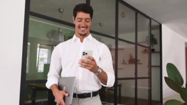 Yakışıklı, başarılı İspanyol erkek girişimci. Kapalı alanda akıllı telefon kullanıyor, koridorda dizüstü bilgisayar taşıyor, telefonda gülümseyen iş adamı mesajları yazıyor.