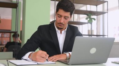 Odaklanmış ofis çalışanı kalemle dizüstü bilgisayarın önünde oturuyor, araştırma yapıyor, webinar izliyor ve notlar alıyor.