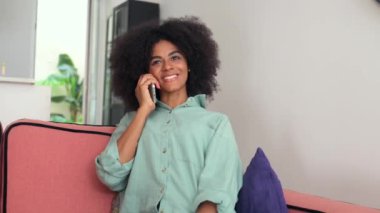Happy african-american woman chatting on the smartphone sitting on the sofa at home, enjoying pleasant phone conversation, gossiping with best friend, smiling