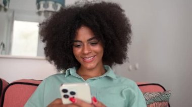 Calm and carefree african lady sitting in a relaxed pose on the comfortable couch with a smartphone, woman spends leisure time scrolling news feed, web surfing, texting with friends