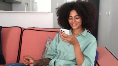 Young african woman sending voice message, recording note through smartphone sitting on the sofa in cozy modern apartment at home, using voice recognize feature