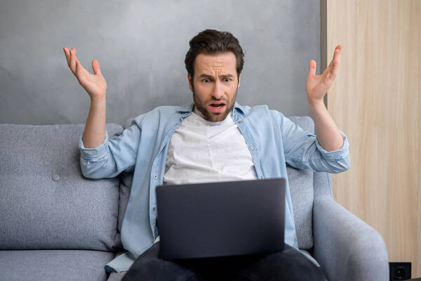 Young hipster guy sitting at the sofa in front of laptop and raising his hands up, feeling frustrated and scared from occurred issues. Businessman is upset to hear the unexpected bad news