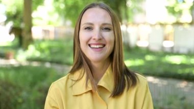 Sıradan gömlekli, gülümseyen ve parkta duran kameraya bakan güzel bir genç kadının portresi. Gülümseyen bir kız öğrencinin açık hava portresi.