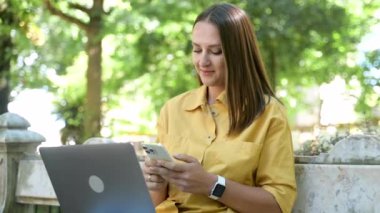 Sarı günlük tişört giyen, akıllı telefondan mesaj atan, dizüstü bilgisayar kullanan, bankta oturan, sosyal ağlarda sohbet eden serbest çalışan genç bir kız öğrenci.