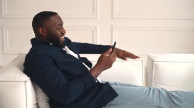 Mutlu 30 'lu yaşlardaki Afrikalı adam sanal buluşmadan zevk alıyor, neşeli mutlu siyah adam akıllı telefondan video görüşmesi yapıyor, evdeki kanepede uzanıyor, uzaktan arkadaşlarıyla ya da ailesiyle irtibatta oluyor.