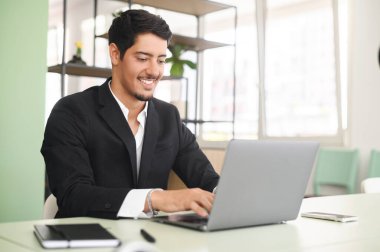 Optimistic ethnic man in formal business wear using laptop sitting on the workplace in modern open space office, young hispanic male employee replying emails, working on a new project
