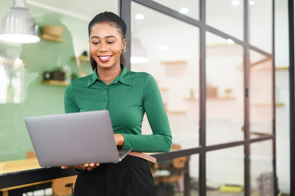 Smart and intelligent African-American businesswoman wearing smart casual shirt using laptop indoors, female office employee holding computer and typing message, smiling, enjoying her job