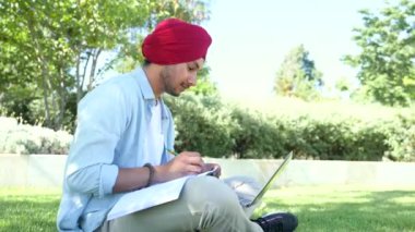 E-öğrenme. Başlıklı çok ırklı Hintli bir adam online kurslar izlemek için dizüstü bilgisayar kullanıyor, ağlar takıyor, kampüsteki parkta oturmuş, erkek öğrenci sınavlarına hazırlanıyor, eğitim dersleri alıyor.