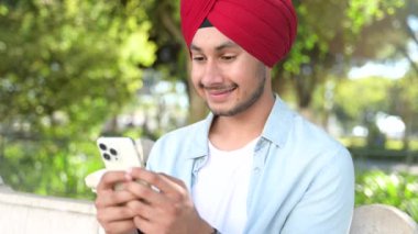 Mutlu ve gülümseyen Hintli adam rahat gömlekli ve kırmızı başlıklı akıllı telefonu açık havada kullanıyor, mesajlaşıyor, sohbet ediyor, internette sörf yapıyor dinlenmiş atmosferde yeni mobil uygulama kullanıyor.