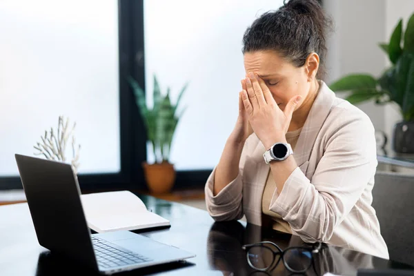 Tired female student feeling bored and tired. Attractive brunette woman sits with eyes closed in front of the laptop, leans head on the hand, feels headache and burnout