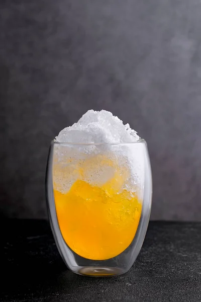 Orange juice with crushed ice on top in glass standing on black table in bar. Fresh delicious beverage made of citrus fruit on grey background close view