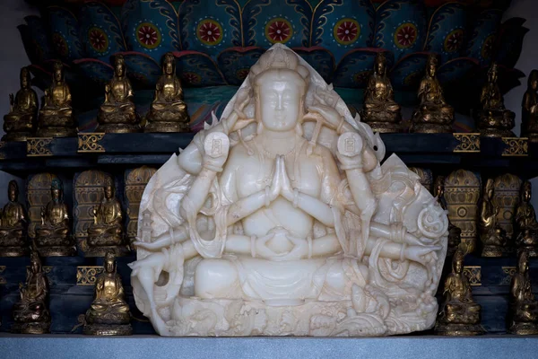 White marble sculpture of Buddha sitting in Lotus pose with hands in prayer gesture on platform against golden statues. Spiritual and religious symbol closeup