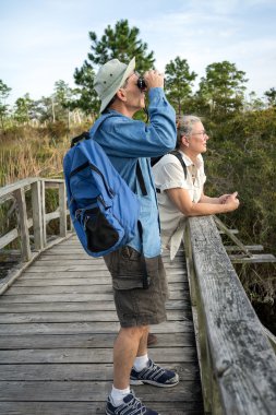 Senior Couple Hiking and Birdwatching on Old Wooden Foot Bridge clipart