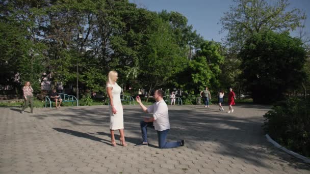 Joven enamorado está de rodillas y hace una oferta con la ayuda de anillo a su amada mujer, multitud de personas corre a la pareja, gritando no en el parque fondo de los árboles — Vídeo de stock