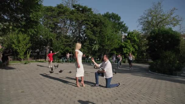 Coppia innamorata, uomo felice in possesso di anello d'oro con diamante in scatola rossa e in ginocchio fa un'offerta alla sua amata donna sfondo di persone che corrono verso lo sfondo coppia di g — Video Stock