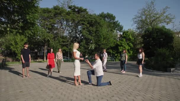 Nowożeńcy, zakochany mężczyzna składa ofertę młodej pięknej kobiecie i kładzie pierścionek na jego palcu w tle grupy ludzi w parku — Wideo stockowe