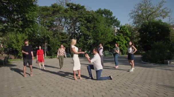 Happy couple, man in love makes an offer to young beautiful woman and puts a ring on his finger backdrop of group of people in park — Stock Video