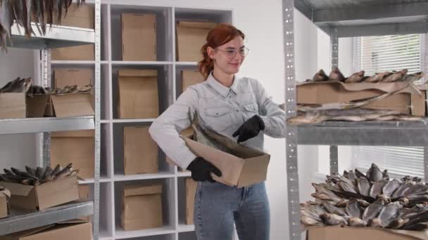 Petite entreprise, souriante jeune femme portant des lunettes ramasse le poisson séché des étagères dans une boîte pour l'envoyer aux clients — Video