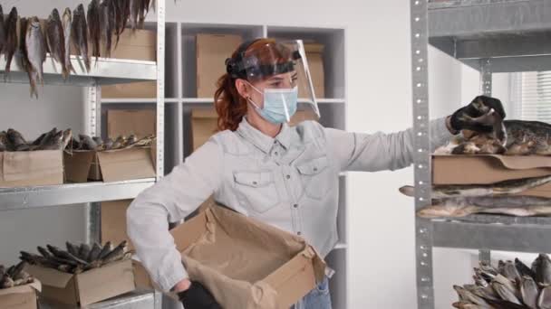 Работа в карантине, женщина в медицинской маске, щит и перчатки ставит соленую рыбу в коробке на заднем плане полок на складе — стоковое видео