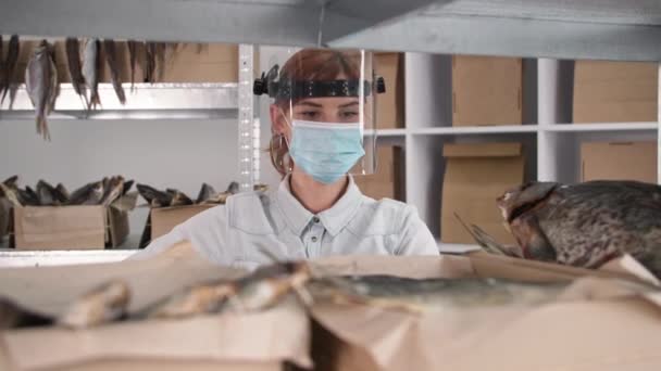 隔離された中小企業医療マスクとシールドを身に着けている若い女性起業家は、オンライン購入者への配達のために箱に魚を入れ — ストック動画