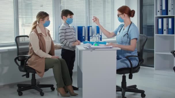 Médico de uniforme azul com estetoscópio conta ao menino e sua mãe sobre a vacina, família e terapeuta em máscaras no consultório do hospital — Vídeo de Stock
