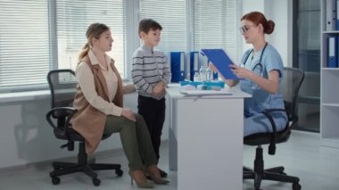 Doktor randevusunda aile, gözlüklü kadın klinisyen, temassız kızılötesi termometresi olan küçük bir hastanın ısısını ölçer ve aşıyı ampulde gösterir.