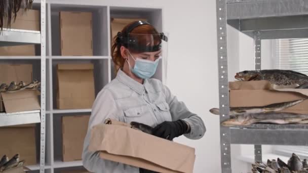 Affärskvinna, ung kvinna i medicinsk mask och visir sätter fisk på hyllor i lager medan du arbetar i karantän — Stockvideo