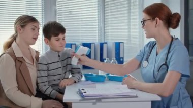 Hasta ve doktor portresi, sağlık görevlisi, çocuk sıcaklığını temas olmayan termometreyle ölçer ve sağlık kliniğindeki aşıdan bahseder.