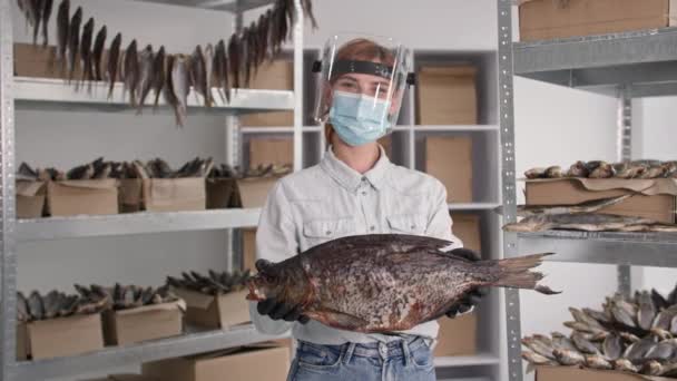 Small business in quarantine, joyful female entrepreneur in medical mask and protective visor with large salted fish in her hands stands backdrop of shelves in warehouse, smiles and looks at camera — Stock Video