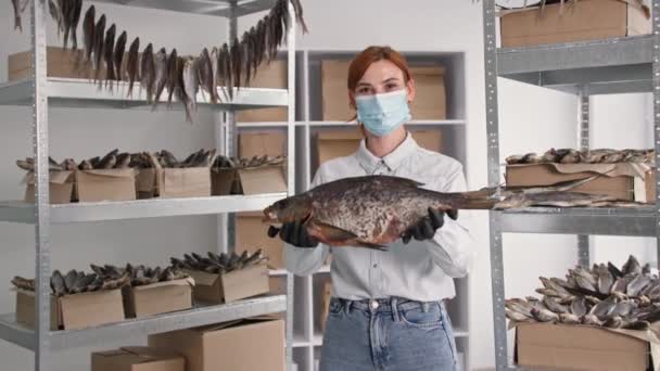 Porträtt av ung kvinna i medicinsk mask med stora havsfiskar i händerna bakgrund hyllor med lådor i lager — Stockvideo