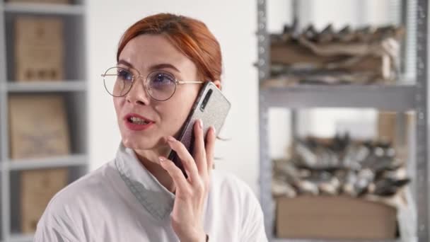 Travail en ligne, jeune femme dans des lunettes parle sur téléphone portable avec le client et prend une commande de poisson séché tout en étant assis dans l'entrepôt sur fond d'étagères — Video