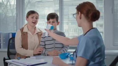 Çocuk ve annesinin doktor randevusundaki portresi. Kadın terapist, temassız termometreyle küçük bir hastanın sıcaklığını ölçer.
