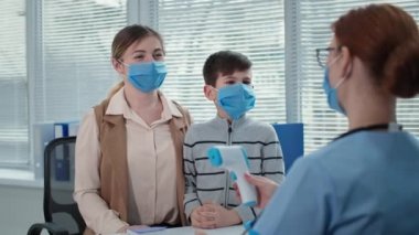 Kadın doktor, annesi hastanede maske takmış bir çocuğa danışıyor. Terapist, temassız termometreyle küçük hastanın ısısını ölçüyor.