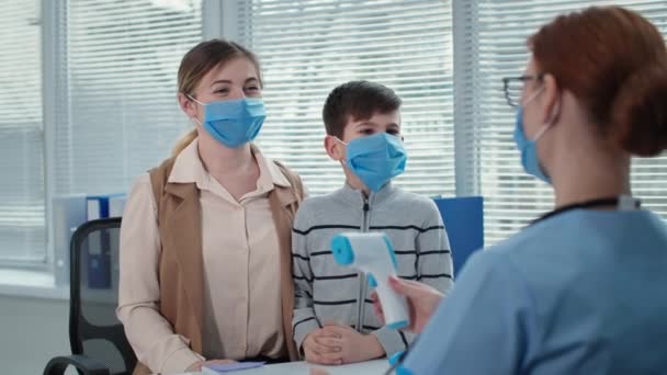 Vrouwelijke arts raadpleegt de jongen met zijn moeder in maskers op afspraak in het ziekenhuis, therapeut meet de temperatuur van de kleine patiënt met een contactloze thermometer — Stockvideo