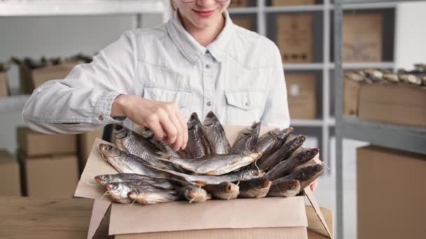 Small business, charming female saleswoman packing dried fish into boxes while sitting on background of shelves in fish store — Stock Video