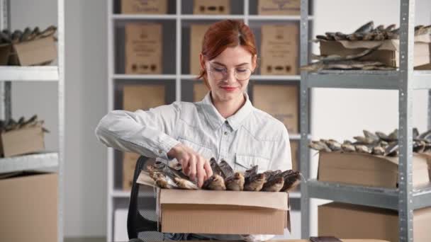 Fish shops, portrait of charming young woman with box of fish in her hands, smiling and looking at camera while sitting in store on background of shelves — Stock Video