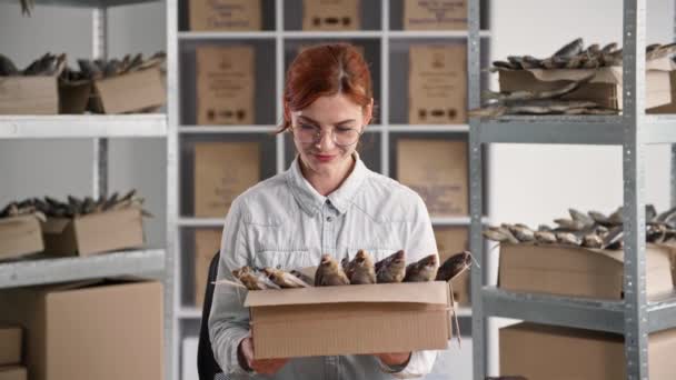 Portrait of beautiful businesswoman with box of dried fish in her hands smiling and looking at camera while sitting in warehouse on background of shelves — Stock Video